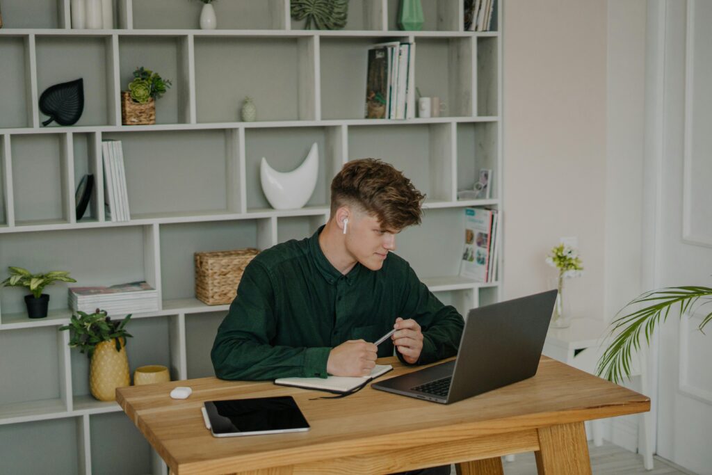 A young man using a laptop for online learning at home, seated at a wooden desk.